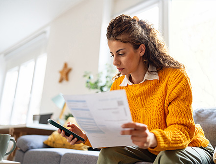 A woman wearing a yellow sweater looks cozy while sitting on the couch in her living room while holding important financial documents and checking her credit history on her cell phone.