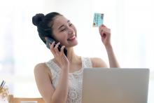 Young woman sitting at desk in front of laptop, using mobile phone, holding a Cathay Bank credit card